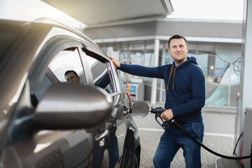 Young cheerful Caucasian man in casual outfit, refueling his modern luxury car at fuel station,...