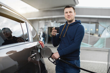 Front view of young Caucasian man, standing outdoors at the gas station, drinking take away coffee...