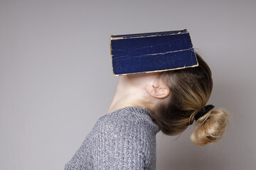  Horizontal image of a young woman covering her face with a book. A beautiful woman in a gray...