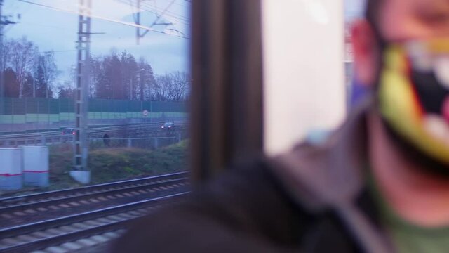 Defocused view of passanger in mask sitting ner window in wagon of train passing by road and rail way.