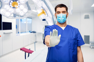 Fototapeta na wymiar healthcare, surgery and medicine concept - doctor or male nurse in blue uniform, face medical mask for protection from virus disease and gloves showing stop gesture over hospital on background