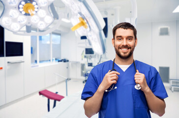 Naklejka na ściany i meble healthcare, surgery and medicine concept - happy smiling doctor or male nurse in blue uniform with stethoscope over operating room at hospital on background