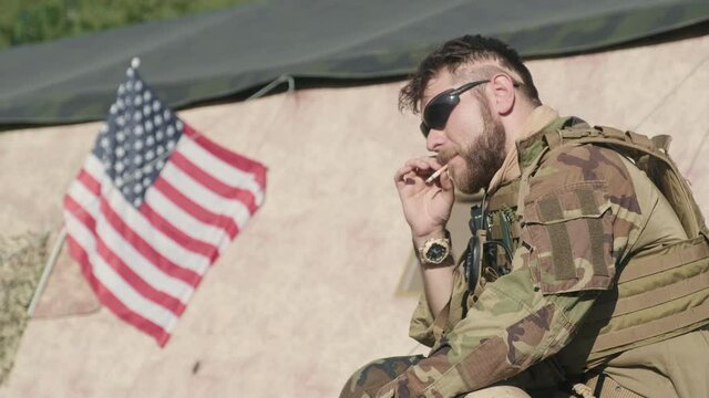 Tilting-up footage of brutal US army officer in camouflage uniform smoking cigarette sitting on army cases outside