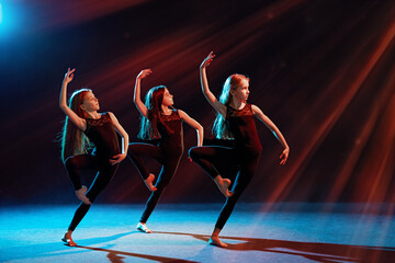 group of three ballet girls in tight-fitting costumes dance against black background with their long hair down, silhouettes illuminated by color sources.