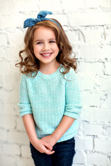 beautiful smiling girl with curly hair in jeans and a blue sweater standing against a white brick wall. Fashion kids