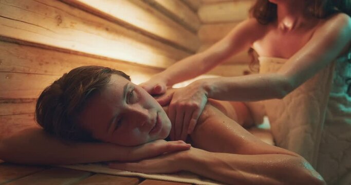 Two women in sauna. Young woman relaxes in a hot wooden sauna and being massaged by her female friend
