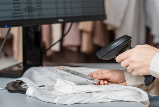 A warehouse woman employee accepts clothes using barcode scanner reading a bar code from price tag of female blouse and adds to the computer base.