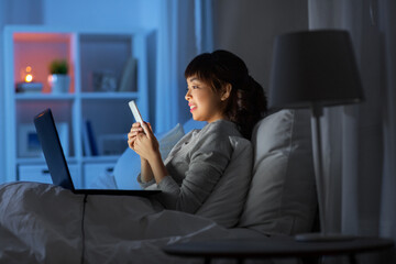 technology, internet, communication and people concept - happy smiling young asian woman with smartphone and laptop lying in bed at home at night