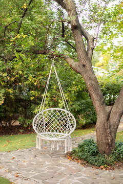 Comfortable hanging wicker white chair in summer garden. Cozy hygge place for weekend relax in garden. Hammock chair in boho style hanging on tree. Cozy exterior backyard. Concept of rest outdoor.	
