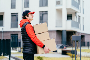 Side view of male in red uniform holding paper boxes for parcels, close-up. Light grey background with apartment house, place to insert your text. Delivery man.