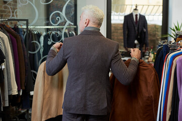 A photo from behind of a mature man with gray hair and a sporty physique is going with two jackets to the fitting room in a clothing store. A male customer with a beard wears a wool suit in a boutique