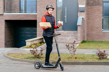 Paper pocket and food containers, box with pizza, disposable coffee cups in hands of a smiling delivery man with kick scooter outdoors. Quality service of a restaurant.