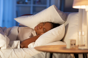 people, bedtime and rest concept - annoyed african american woman lying in bed at night and covering ears with pillow