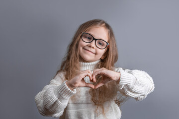 Portrait of a young attractive little girl with blond long flowing hair in a white sweater smiling shows heart hands on a gray studio background. Place for text. Copy space