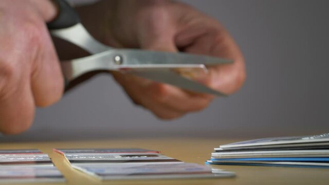 Closeup POV shot of a man’s hands taking a credit or membership card from a pile, then using a pair of scissors to cut it in half. Other cut cards lie nearby. Money trouble concept.