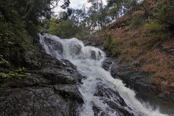 Fototapeta premium The waterfall cascades down from the rocks. Splashes and foam over water stream. Green plants all around. Vietnam. Dalat