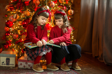 Cute twins girls in red sweaters read a book sitting near boxes with presents near a Christmas tree in a decorated room