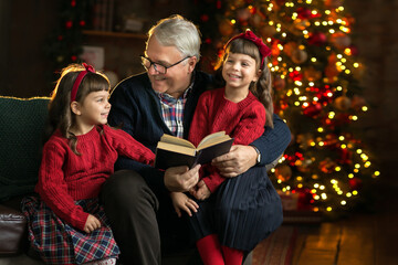 Grandfather wearing glasses, reading a book to small granddaughters twins in a room decorated for Christmas on the background of a Christmas tree. Christmas holiday concept.