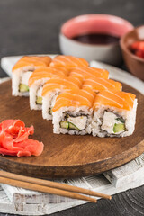 Classic Philadelphia sushi rolls on a wooden board with ginger, soy sauce and chopsticks on a dark background.