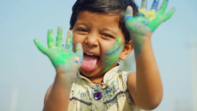young cute little girl kid with applied holi colors powder dancing with grimacing by showing colorful hands to camera