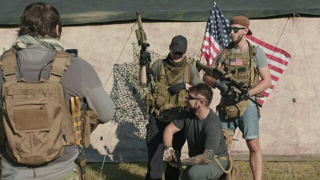 Medium shot of young military men in US army taking photo with weapons posing for camera with US flag flattering in background