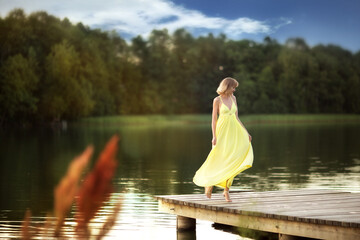 beautiful young blond woman in a yellow dress dancing on a pier at the river