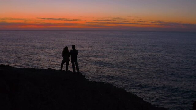 Silhouette of unrecognisable couple stand on edge of cliff, watch over sunset or sunrise, sun behind horizon. Cinematic and fading to black video of adventurers rise early on mountain peak