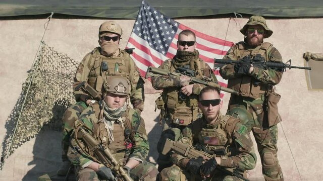 Medium portrait of young US intelligence officers in camouflage standing together holding rifles with US flag flattering in background