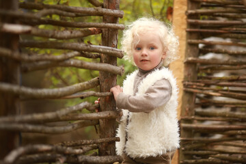 Cheerful girl with curly white hair in a vest standing near a fence in a spring garden