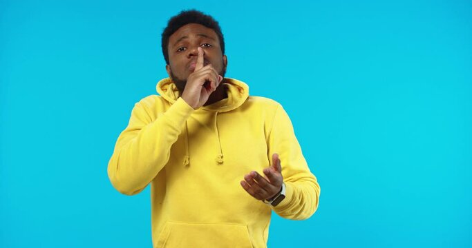 Close up portrait of serious African American young male shrugs waving his finger saying NO rejecting something disagreeing making blah gesture with hands standing isolated on blue wall, be quiet