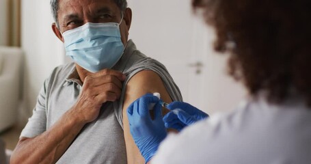 Mixed race female doctor wearing mask giving vaccination to senior man at home - Powered by Adobe