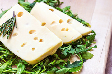 Maasdam cheese slices with green arugula leaves on light wooden background.
