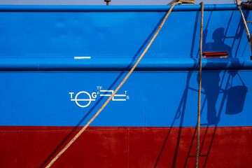 Sunlight and shadow of painter climbing ladder with white plimsoll mark on blue and red rust proof steel hull surface of fishing vessel with mooring rope in shipyard area