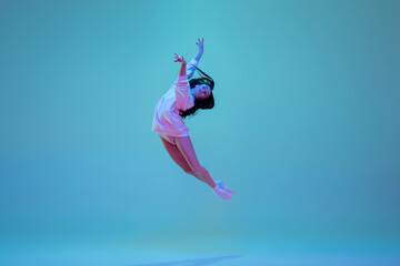 Flying high. Young and graceful ballet dancer isolated on blue studio background in neon light. Art, motion, action, flexibility, inspiration concept. Flexible caucasian ballet dancer, moves in glow.