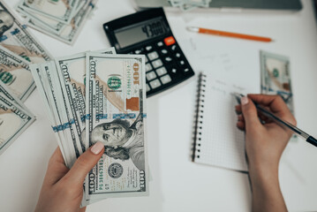 Unrecognizable woman holding money in hands and calculates income on calculator. Tracking home budget and expenses. Accountant or banker working - top view.