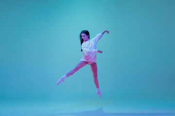 Fototapeta na wymiar In flight. Young and graceful ballet dancer isolated on blue studio background in neon light. Art, motion, action, flexibility, inspiration concept. Flexible caucasian ballet dancer, moves in glow.