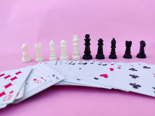 White and black Chess pieces and deck of playing cards facing each other. Pink background