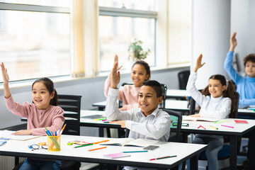 Diverse small schoolkids raising hands at classroom