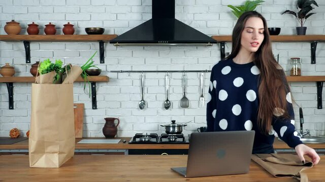 Woman chef teacher comes to home kitchen puts on beige apron prepare teaches online culinary webinar course in laptop computer, paper bag with fresh food vegetables for cooking dish stands on table