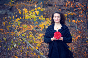  sad brunette woman in black dress holding red flower in forest