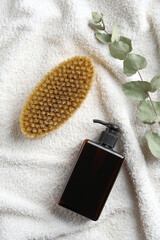 Dry brush for body massage, gel dispenser and eucalyptus leaf on white towel in bathroom. Flat lay, top view.