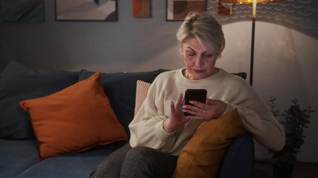 Senior woman using mobile app sitting on sofa, texting typing message hold smart phone look at cellphone screen browsing social media at home, businesswoman tired after work