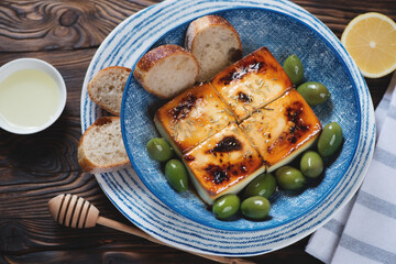 Blue bowl with baked feta cheese, green olives, baguette and olive oil, elevated view on a brown...