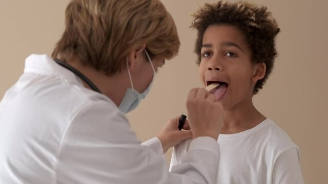 Female practitioner doctor in white coat protective face mask holding medical spatula makes little african boy larynx check up. General health exam, podiatry, laryngoscopy, healthcare concept