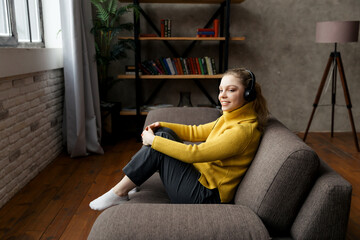 Fototapeta na wymiar Beautiful pleased woman listening to music in headphones while sitting on sofa at home