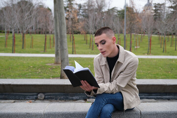 Obraz premium Handsome young man wearing make up, reading a book sitting on a wall. Non binary androgynous guy.