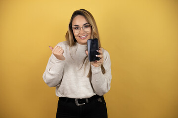 Obraz premium Pretty woman with long hair holding smartphone showing blank screen smiling happy pointing with hand and finger to the side against yellow wall