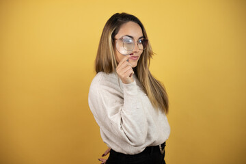 Pretty woman with long hair looking through a magnifying glass against yellow wall