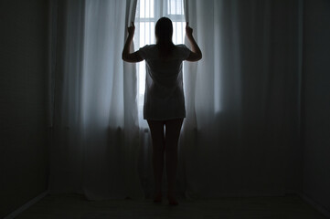 Fototapeta na wymiar A full-length Caucasian woman standing by window in the twilight in morning opens curtains.