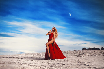 Fototapeta premium A gorgeous blonde girl in a sexy red dress with deep slits on the hips against the background of a white sandy beach and a sunset sky.